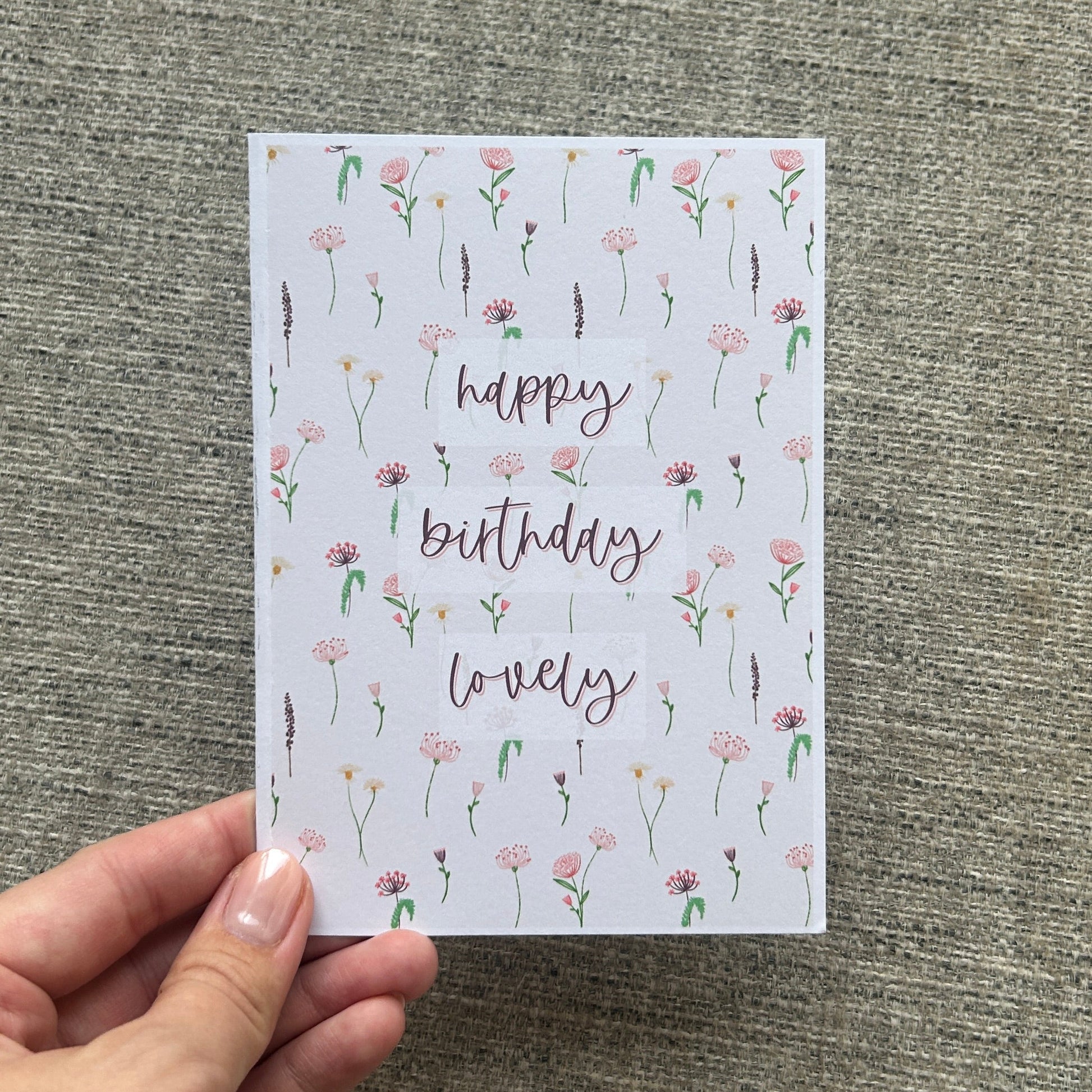 A hand holding a square greeting card with a white background, decorated with a delicate pink and green floral pattern and the words 'happy birthday lovely' in a cursive font, set against a textured gray fabric background.
