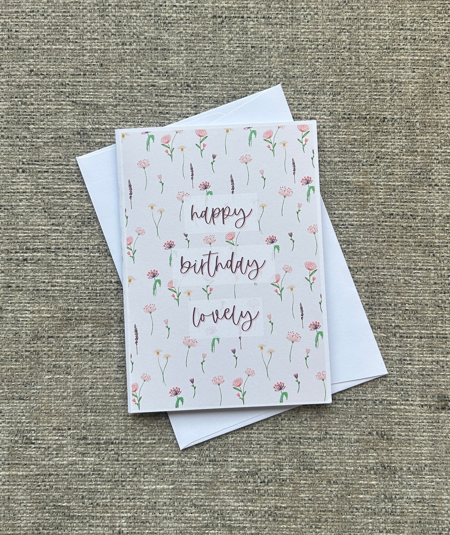 A square greeting card with a white background, decorated with delicate pink and green wildflower illustrations and the words 'happy birthday lovely' in a cursive font. The card is shown with two white envelopes on a textured, woven beige fabric background.