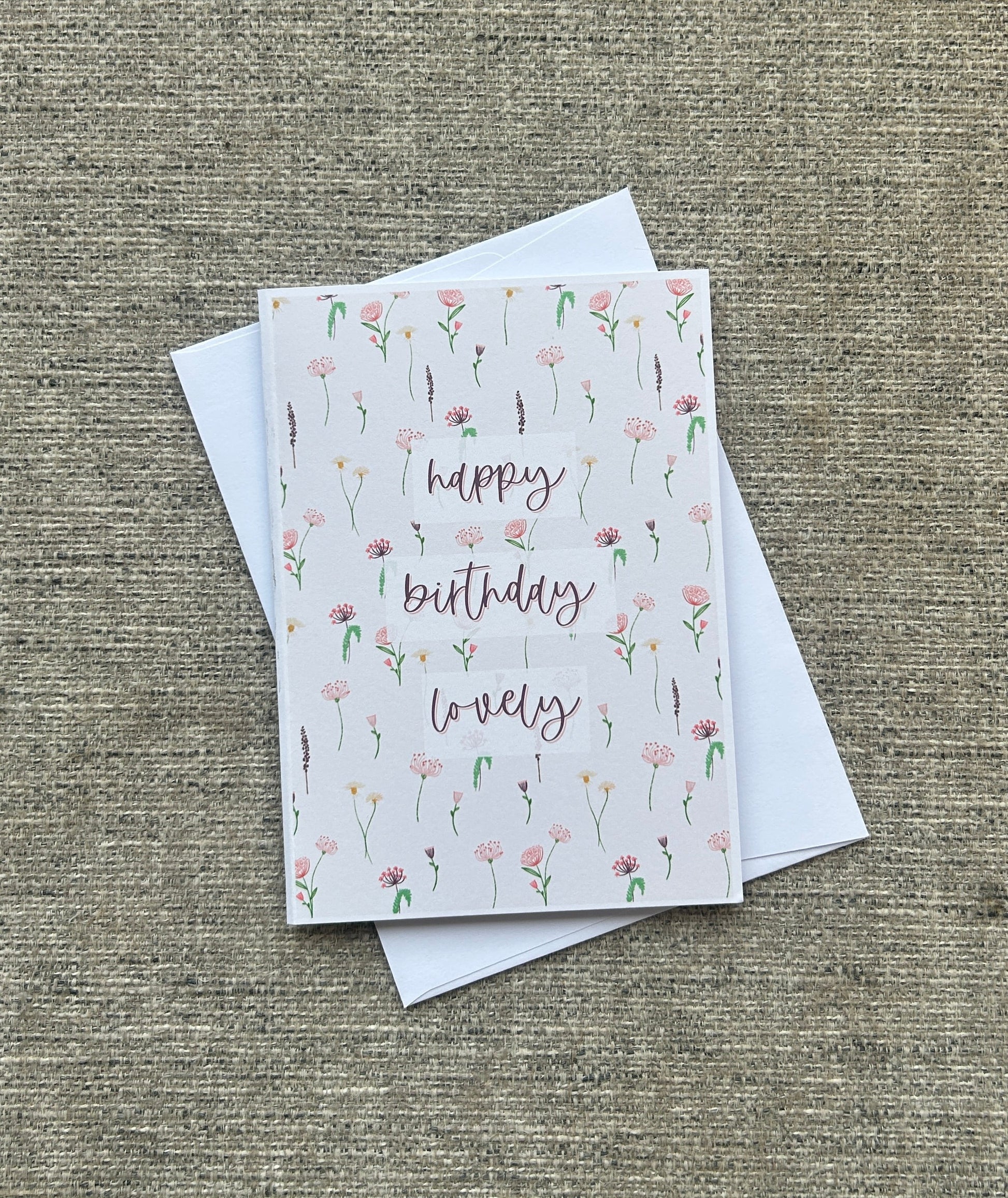 A square greeting card with a white background, decorated with delicate pink and green wildflower illustrations and the words 'happy birthday lovely' in a cursive font. The card is shown with two white envelopes on a textured, woven beige fabric background.