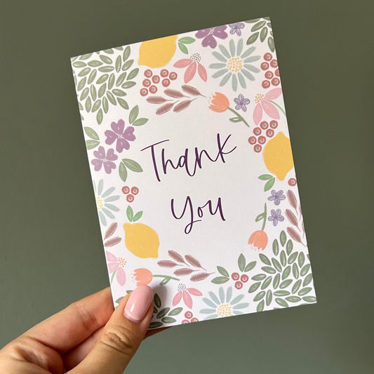 Hand holding a 'Thank You' card with floral design on a plain background