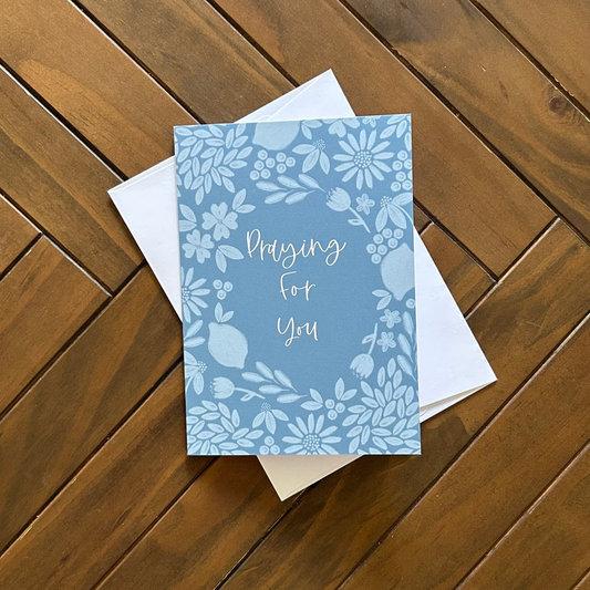 Greeting card with floral design on a blue background and text to say Praying for you on a wooden surface