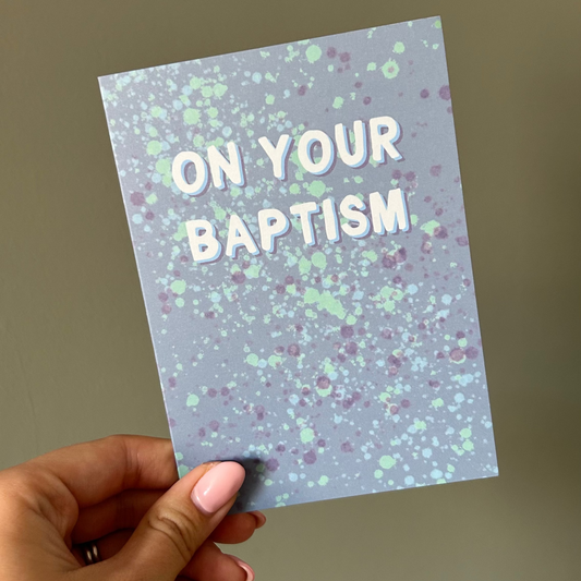 Hand holding a card with 'On Your Baptism' text against a plain background