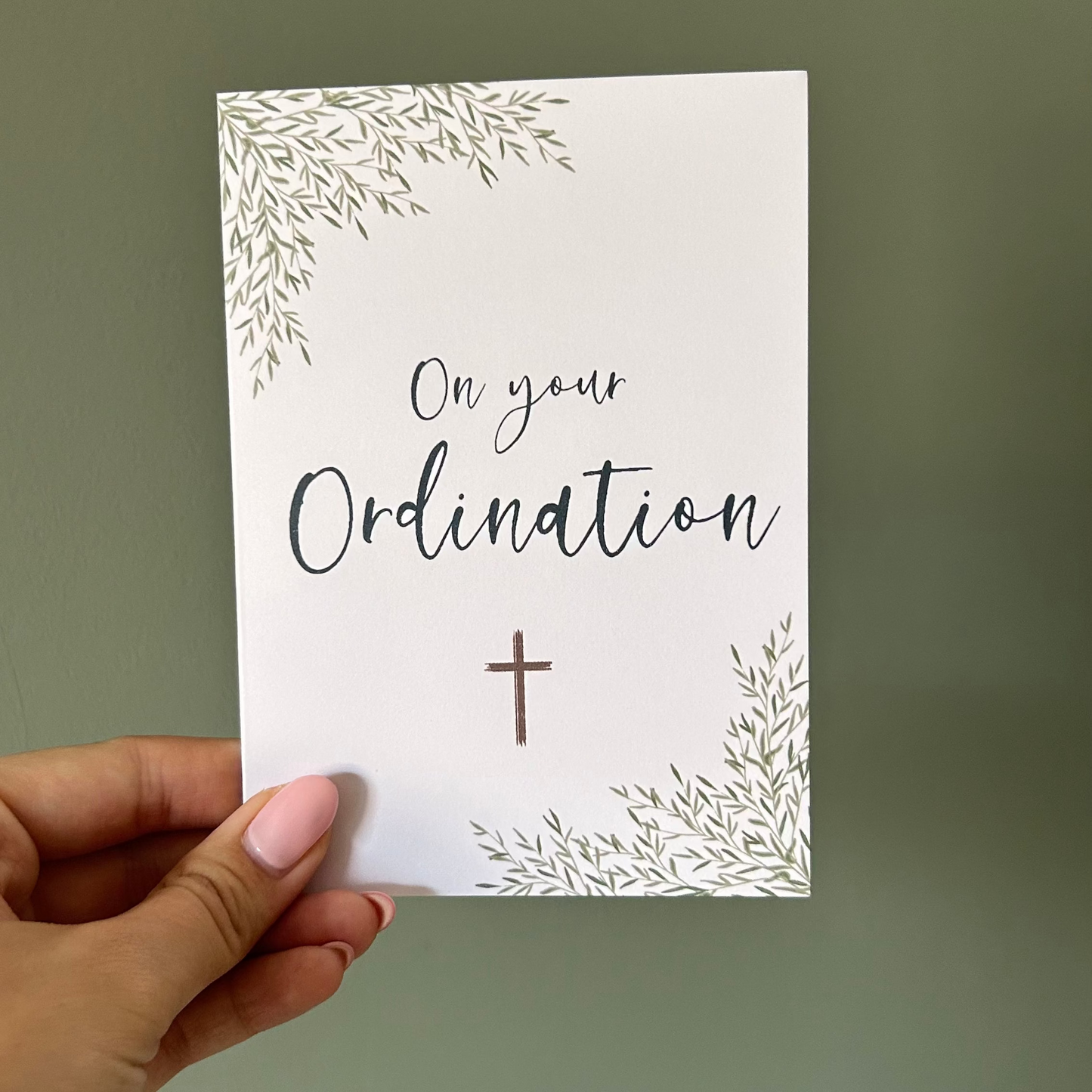 Hand holding a card with 'On your Ordination' text and a cross on a plain background