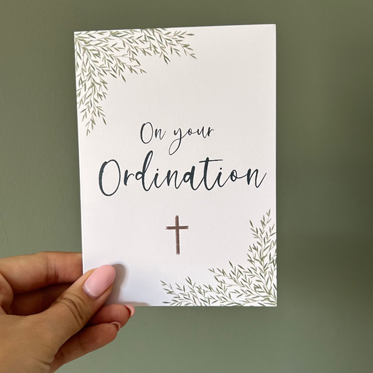 Hand holding a card with 'On your Ordination' text and a cross on a plain background