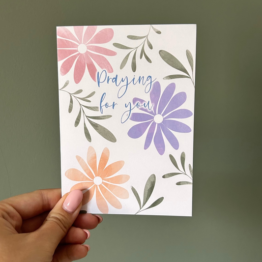 Hand holding a card with floral designs and 'Praying for you' text against a plain background