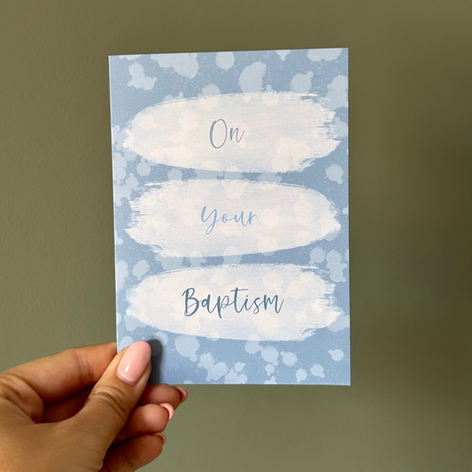 Hand holding a blue baptism card with white text against a plain background