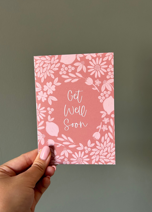 Deep orange floral card with 'Get Well Soon' text held by a hand against a green background