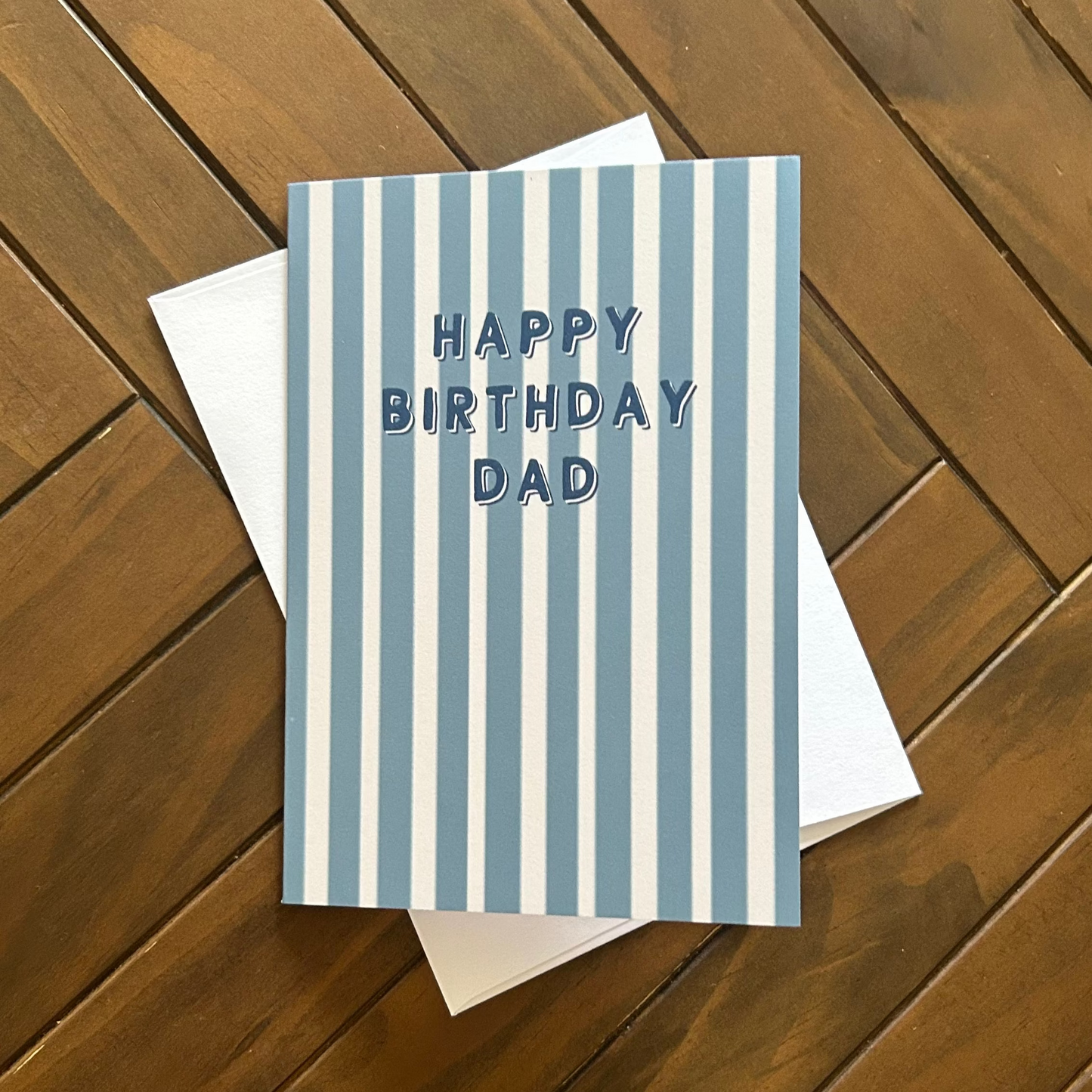 A square birthday card with vertical blue and white stripes and the text 'HAPPY BIRTHDAY DAD' in blue, placed on top of a white envelope over a brown wooden plank surface.