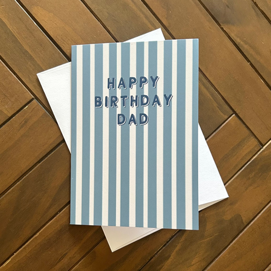 A square birthday card with vertical blue and white stripes and the text 'HAPPY BIRTHDAY DAD' in blue, placed on top of a white envelope over a brown wooden plank surface.