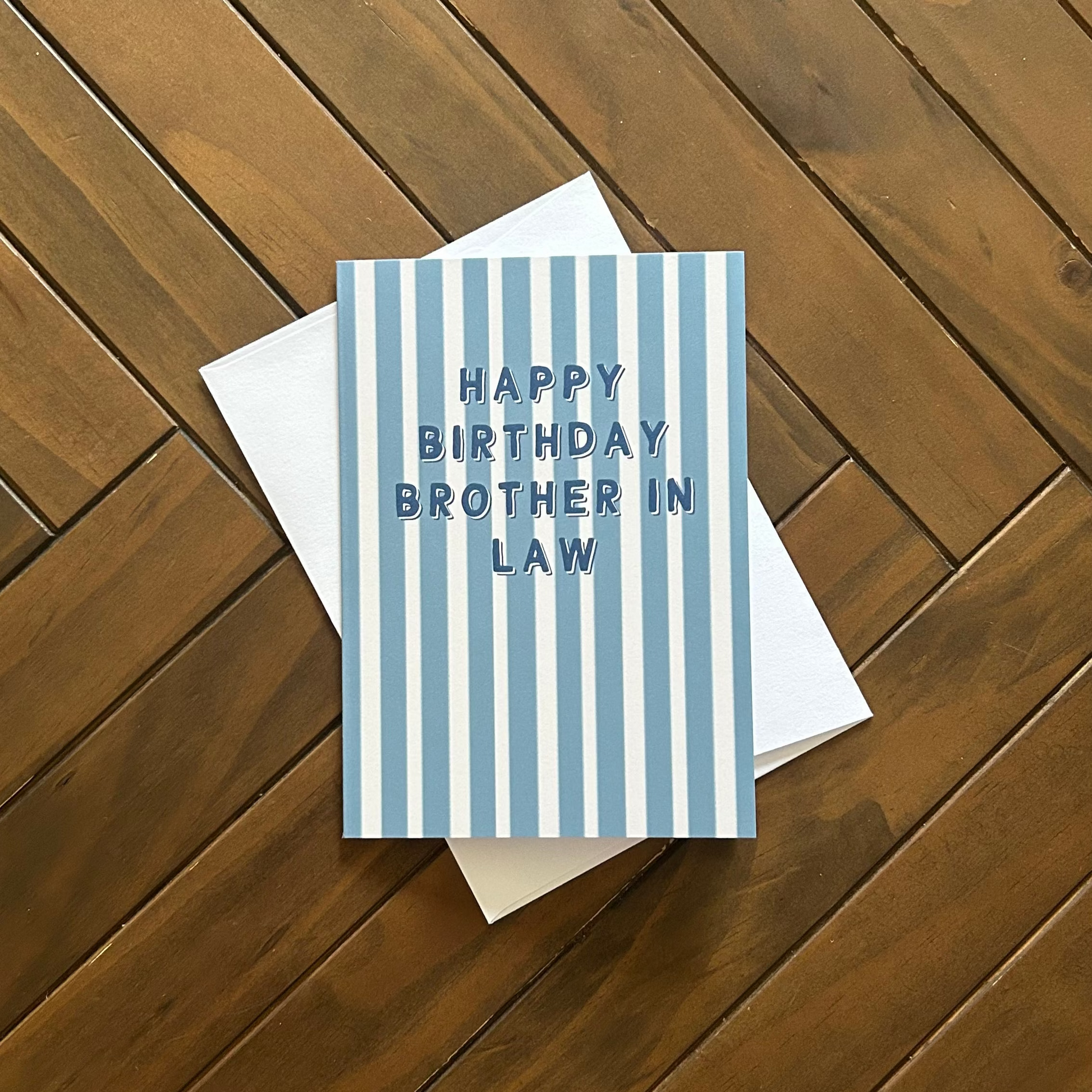 A blue and white striped greeting card with the text 'HAPPY BIRTHDAY BROTHER IN LAW' in navy blue, resting on a white envelope over a brown wooden plank background.