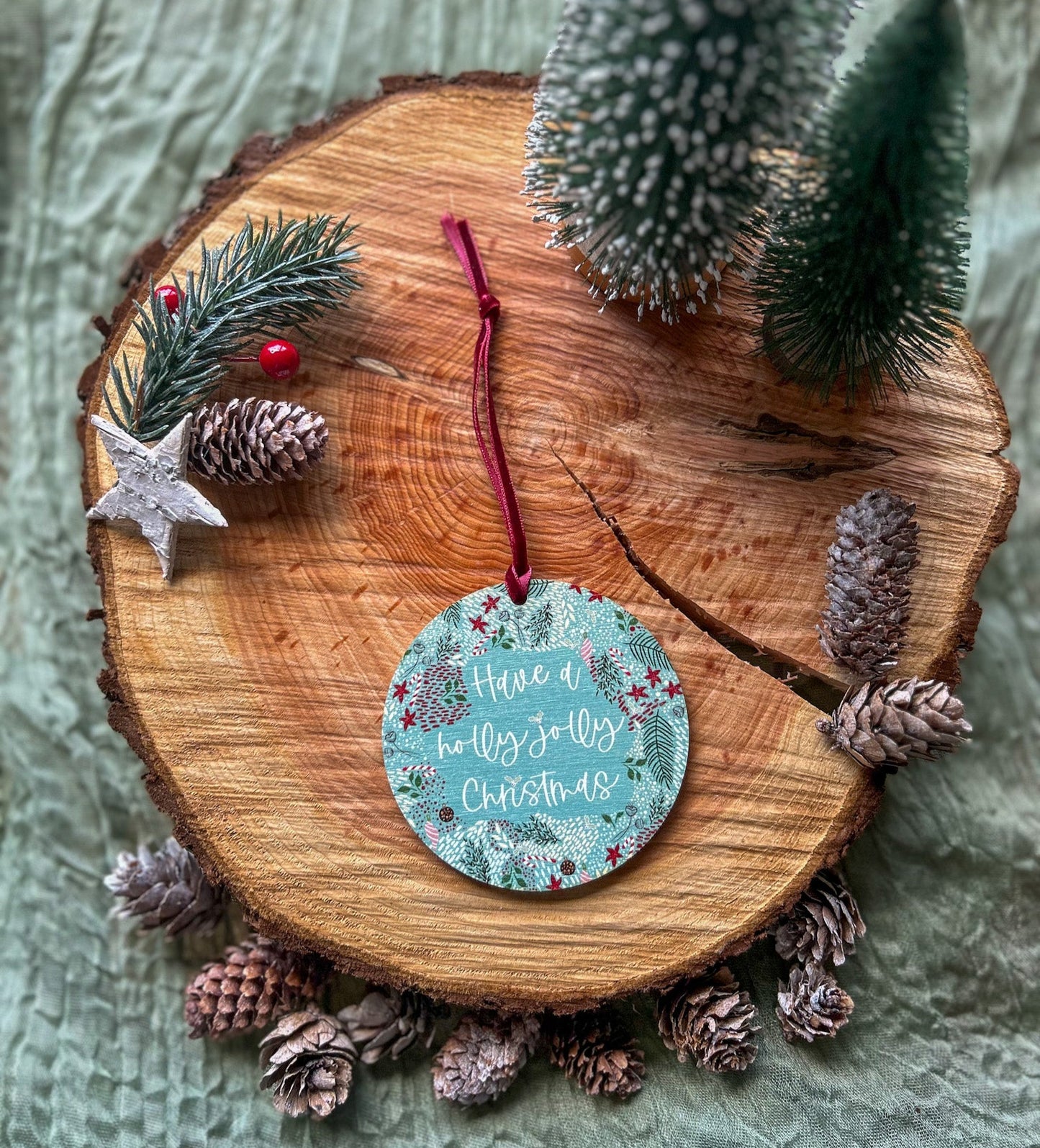 Wooden hanging decorations with 'Have a holly jolly Christmas' on it and a deep red velvet ribbon on a wooden slice with pinecones and a small tree.