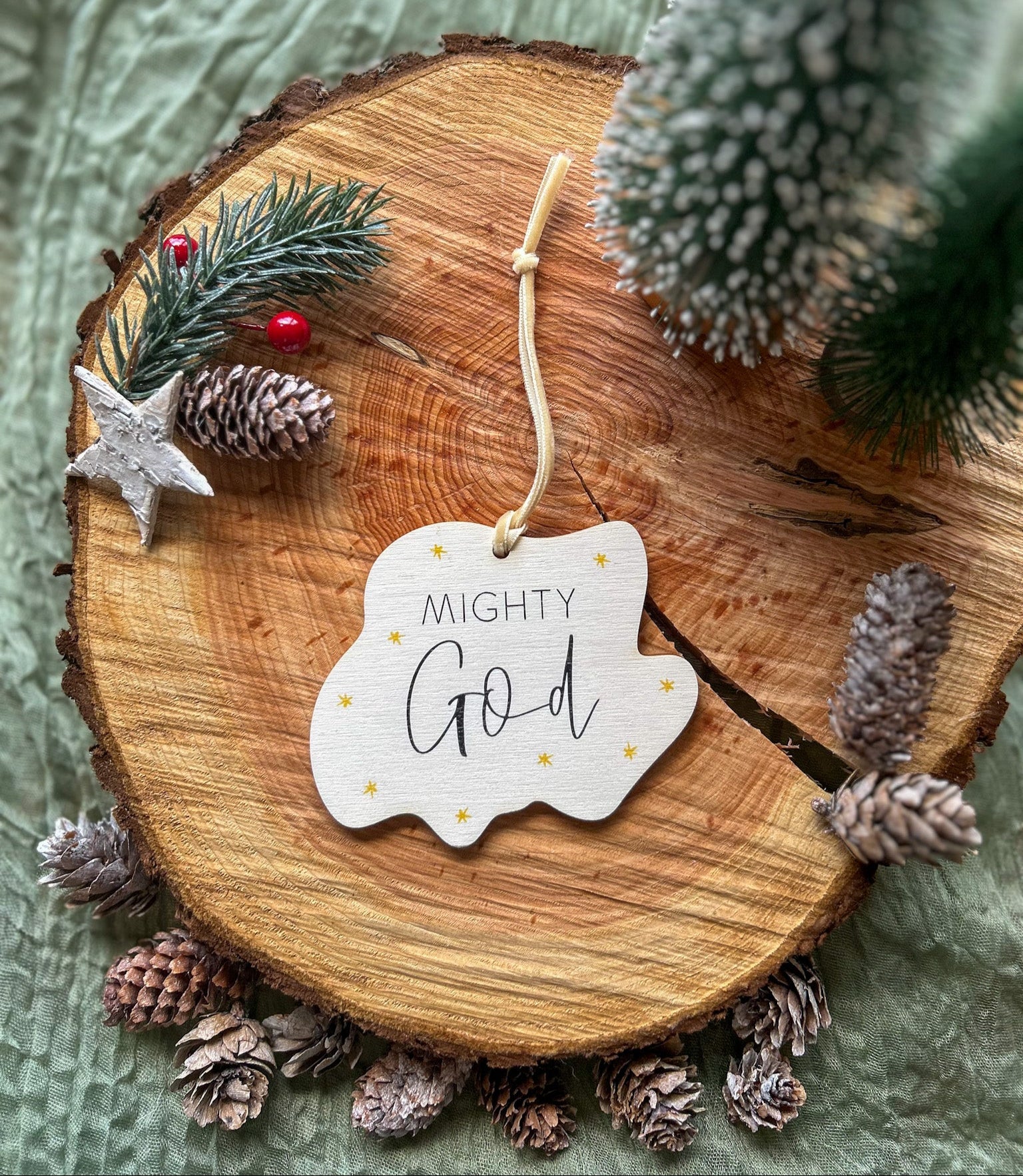 Wooden hanging decoration with 'Mighty God' on it and a creamy gold velvet ribbon on a wooden slice with pinecones and a small tree.