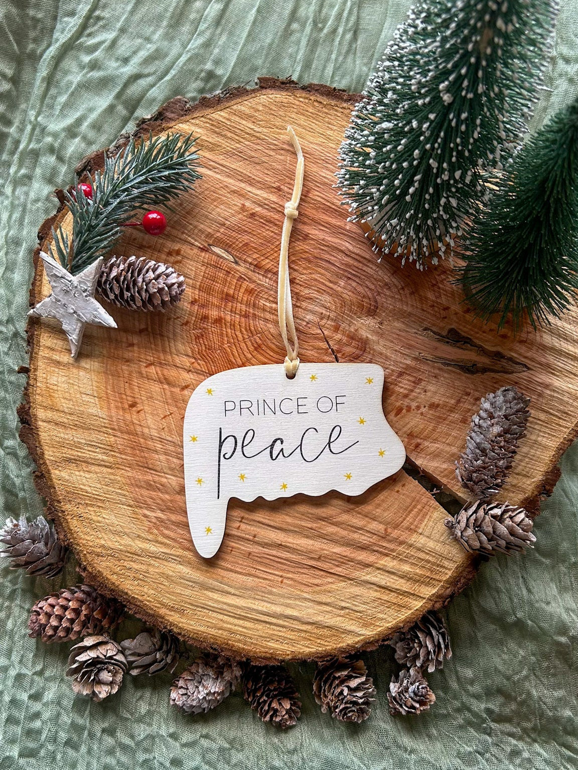 Wooden hanging decoration with 'Prince of Peace' on it and a creamy gold velvet ribbon on a wooden slice with pinecones and a small tree.