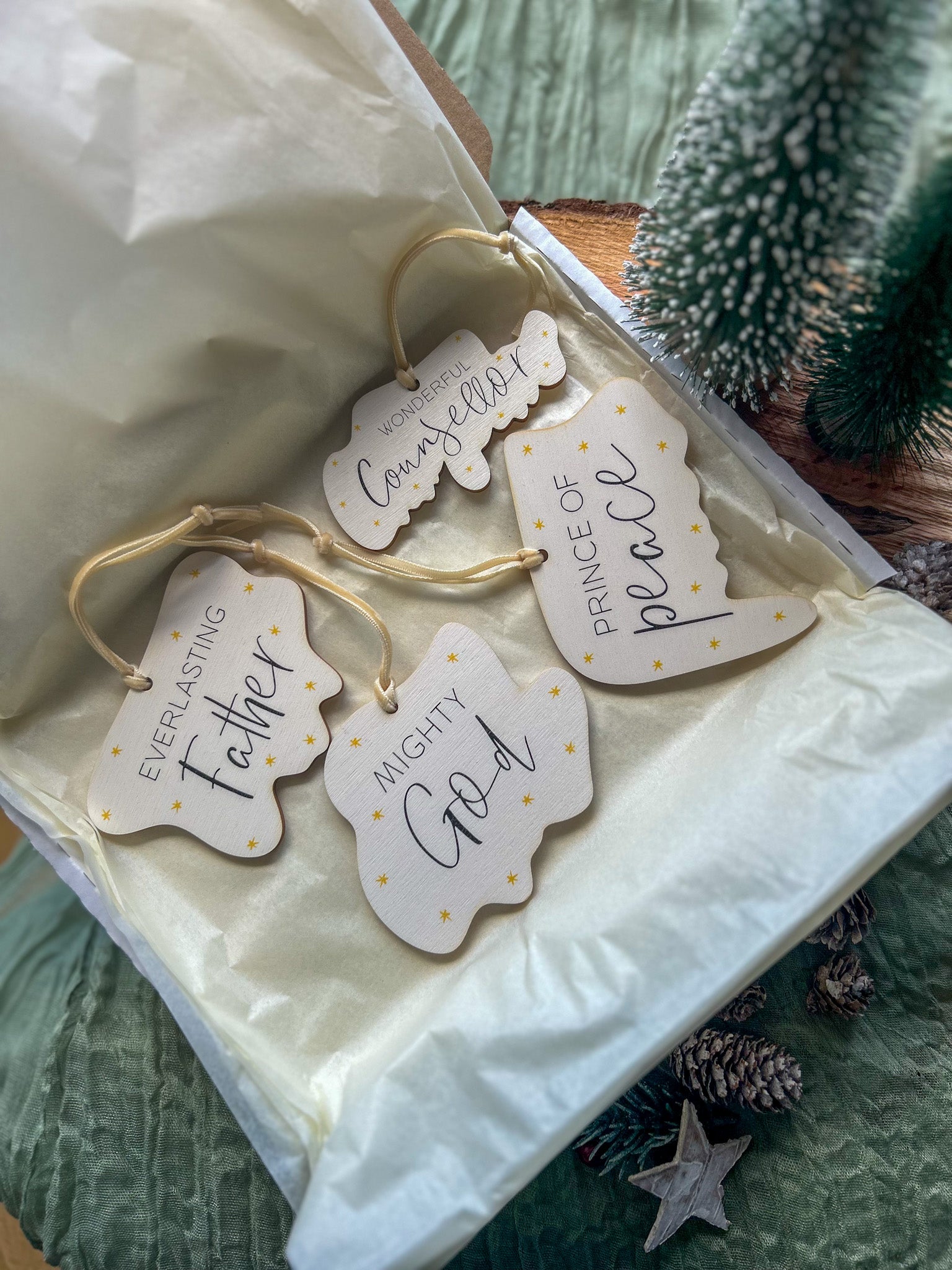 Wooden hanging decorations with 'Wonderful Counsellor', 'Everlasting Father', 'Mighty God' and 'Prince of Peace' on it and a creamy gold velvet ribbon on a wooden slice with pinecones and a small tree.